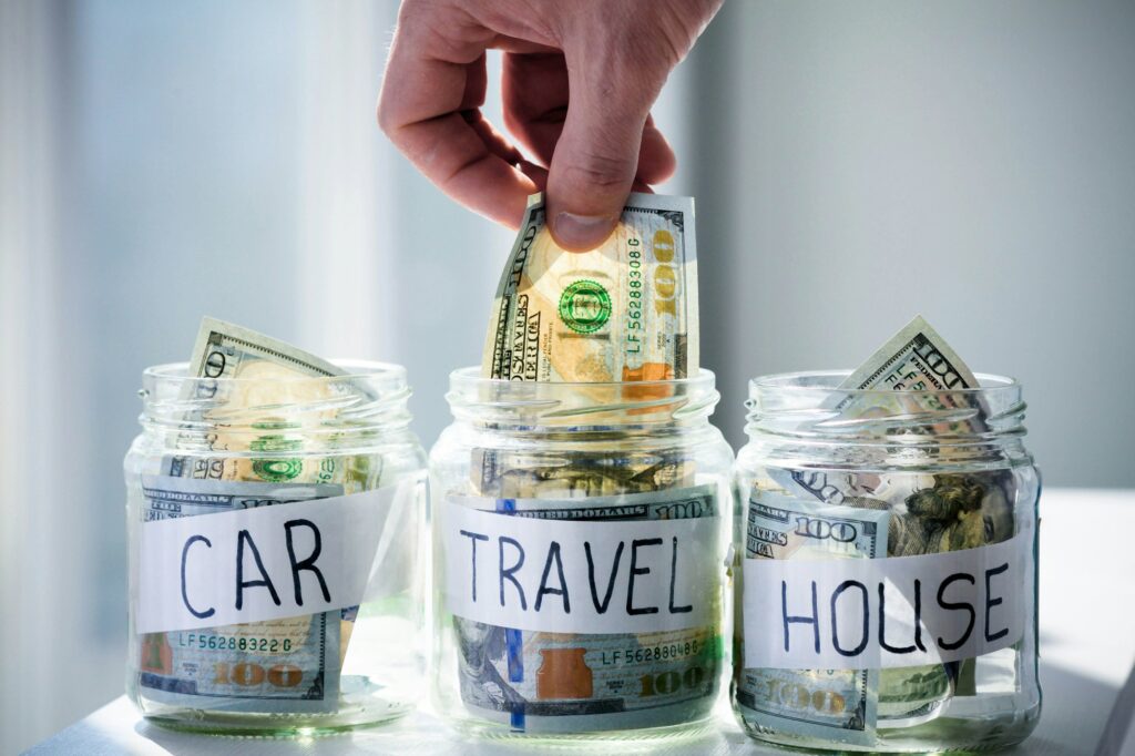 Male hand puts 100 dollars in a glass jar with the inscription travel. - Budgeting for Travel